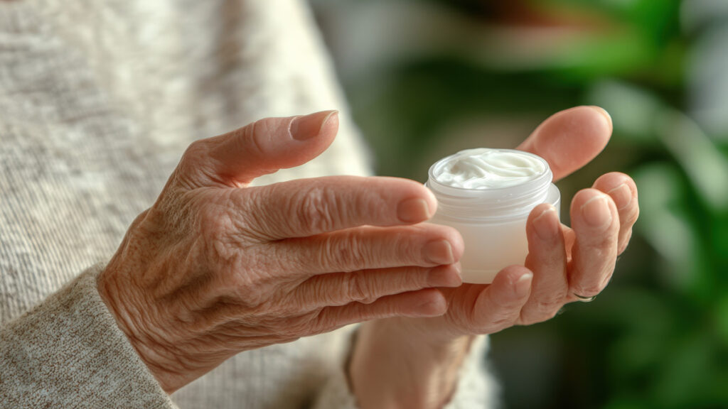 Elderly hands holding a small jar of pain relief cream, ready for application. Blue-Emu helps soothe joint and muscle discomfort.